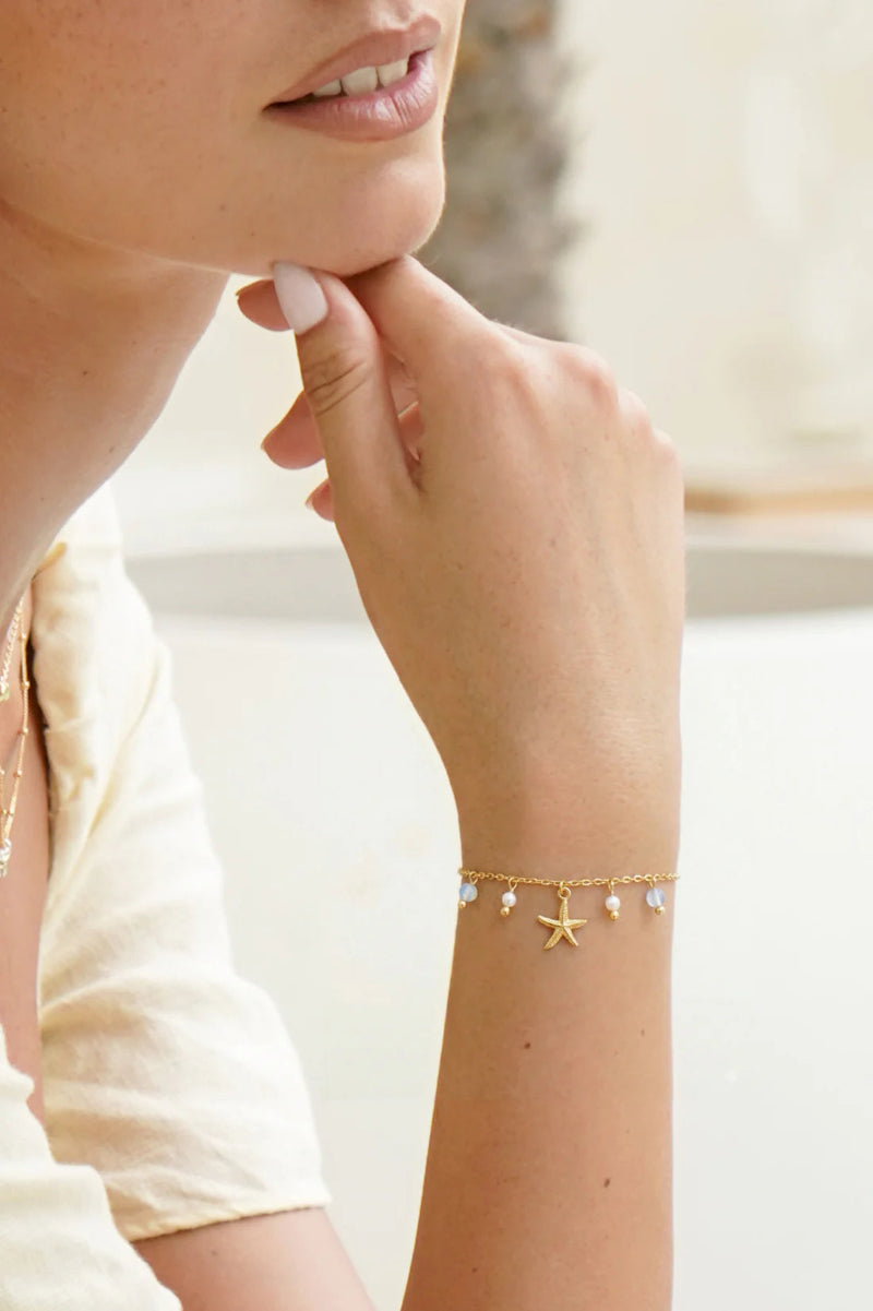 Close-up of a person wearing a gold bracelet with starfish charm on a blurred background