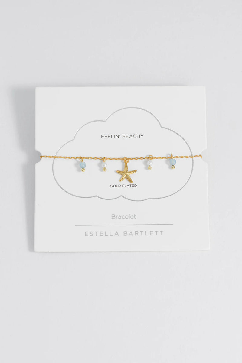 Gold-plated bracelet with star and shell charms on Estella Bartlett packaging.