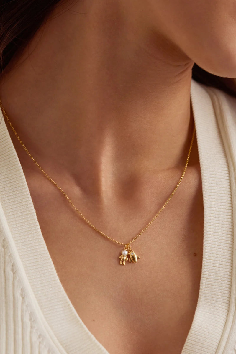 A close-up of a model wearing a delicate gold chain necklace with a small gold and pearl pendant, styled with a cream-colored ribbed knit top.