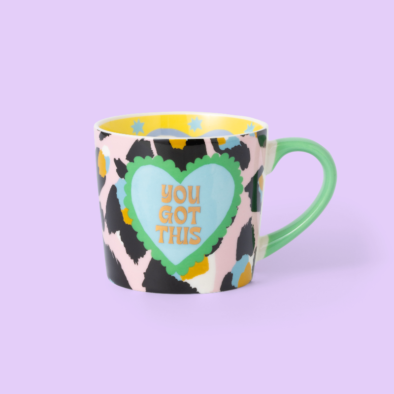 A ceramic coffee mug with a colorful abstract pattern in black, pink, yellow, and blue, featuring a central light blue heart with the text 'YOU GOT THIS' in gold. The mug has a mint green handle and a yellow interior, set against a solid lavender background.