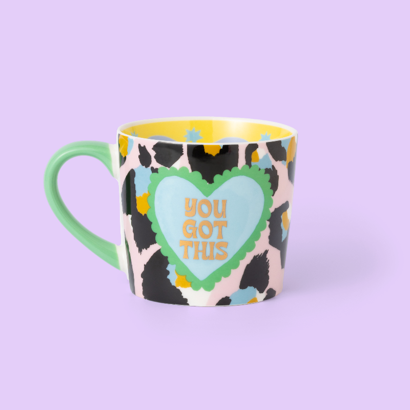 A ceramic coffee mug with a black and beige cow print pattern, featuring a light blue heart in the center with the text 'YOU GOT THIS' in gold. The mug has a mint green handle and a yellow interior, set against a solid lavender background.