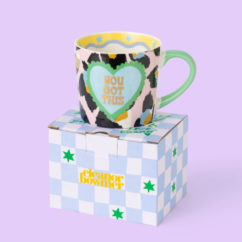 A ceramic mug with a cow-print pattern, a green handle, and a light blue heart graphic that reads 'YOU GOT THIS', displayed on top of a light blue and white checkered box with green stars and yellow 'cleaner bowtie' branding, against a solid lavender background.