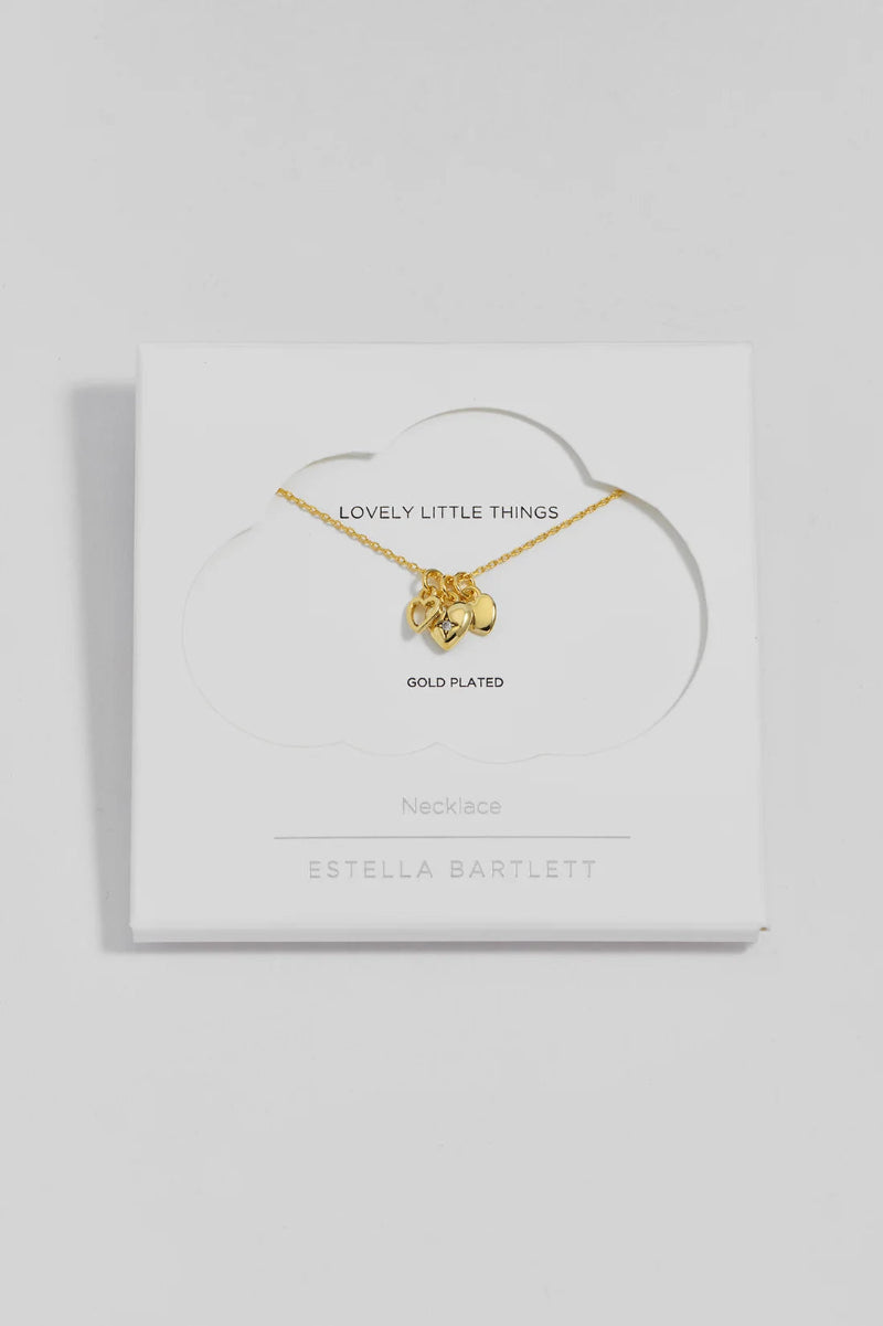 A gold-plated necklace with a small heart charms displayed on a white cloud-shaped card inside a square white box. The card reads 'LOVELY LITTLE THINGS' and 'ESTELLA BARTLETT'.