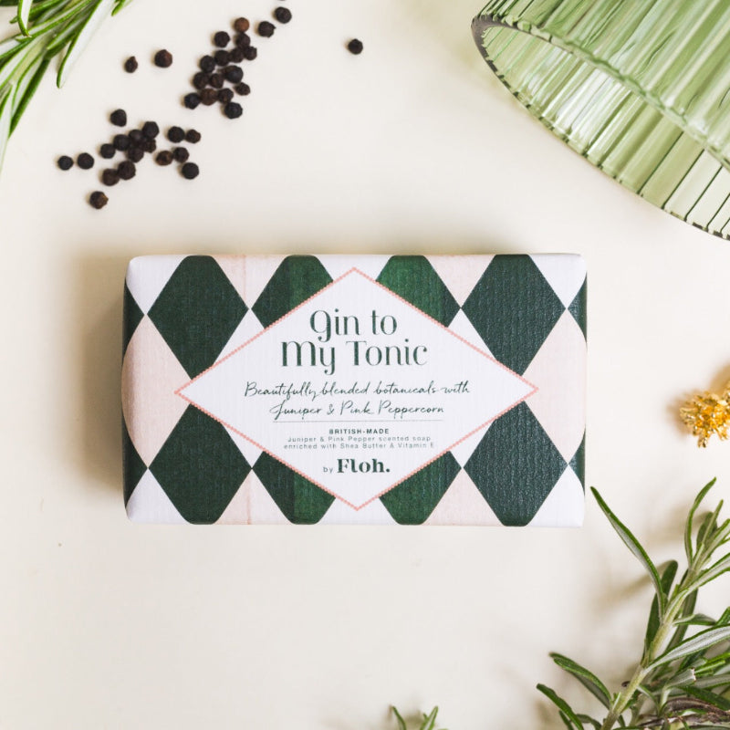 A rectangular bar of soap wrapped in a green and white harlequin patterned paper sleeve, labeled 'Gin to Tonic' by Floh. The soap is styled in a flat-lay arrangement with rosemary sprigs, whole black peppercorns, and a green glass plate on a warm cream background.