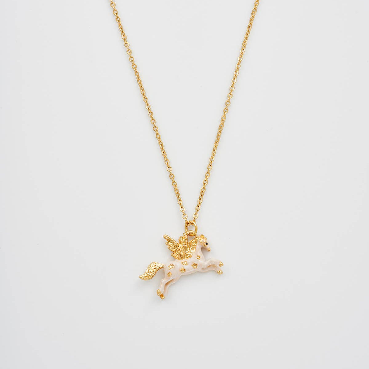 A gold chain necklace featuring a small winged unicorn pendant with pale pink enamel and tiny gold star details, shown against a plain light gray background.