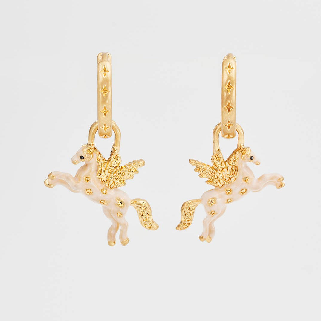 A pair of gold-tone hoop earrings with dangling winged unicorn charms in a pale pink enamel finish, shown against a plain light gray background.