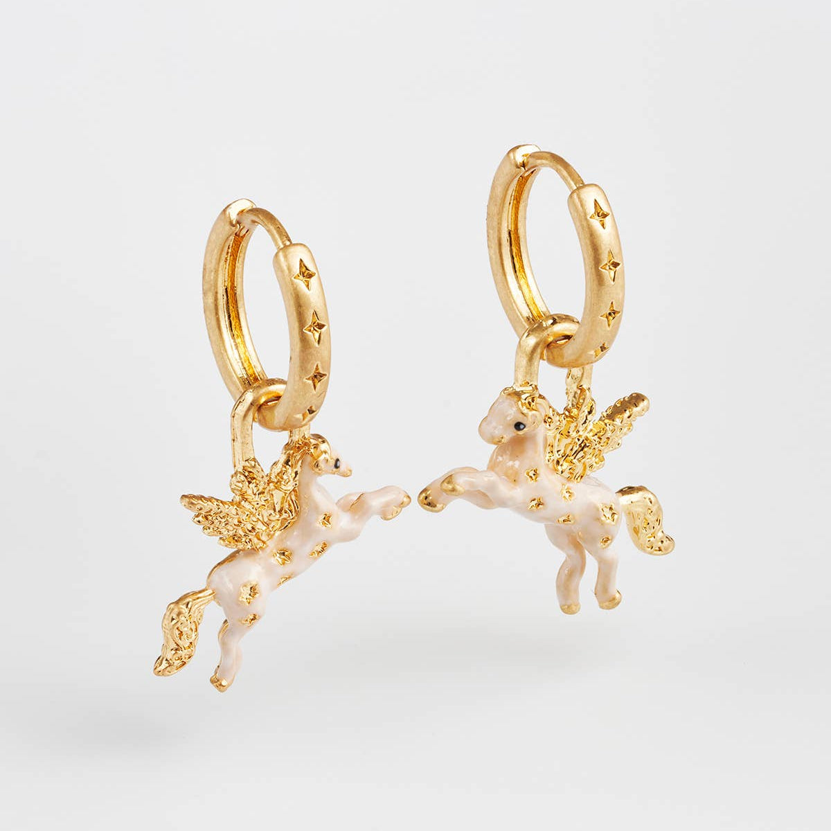 A pair of gold huggie hoop earrings featuring dangling winged unicorn charms with white enamel and gold star details, set against a clean white background.