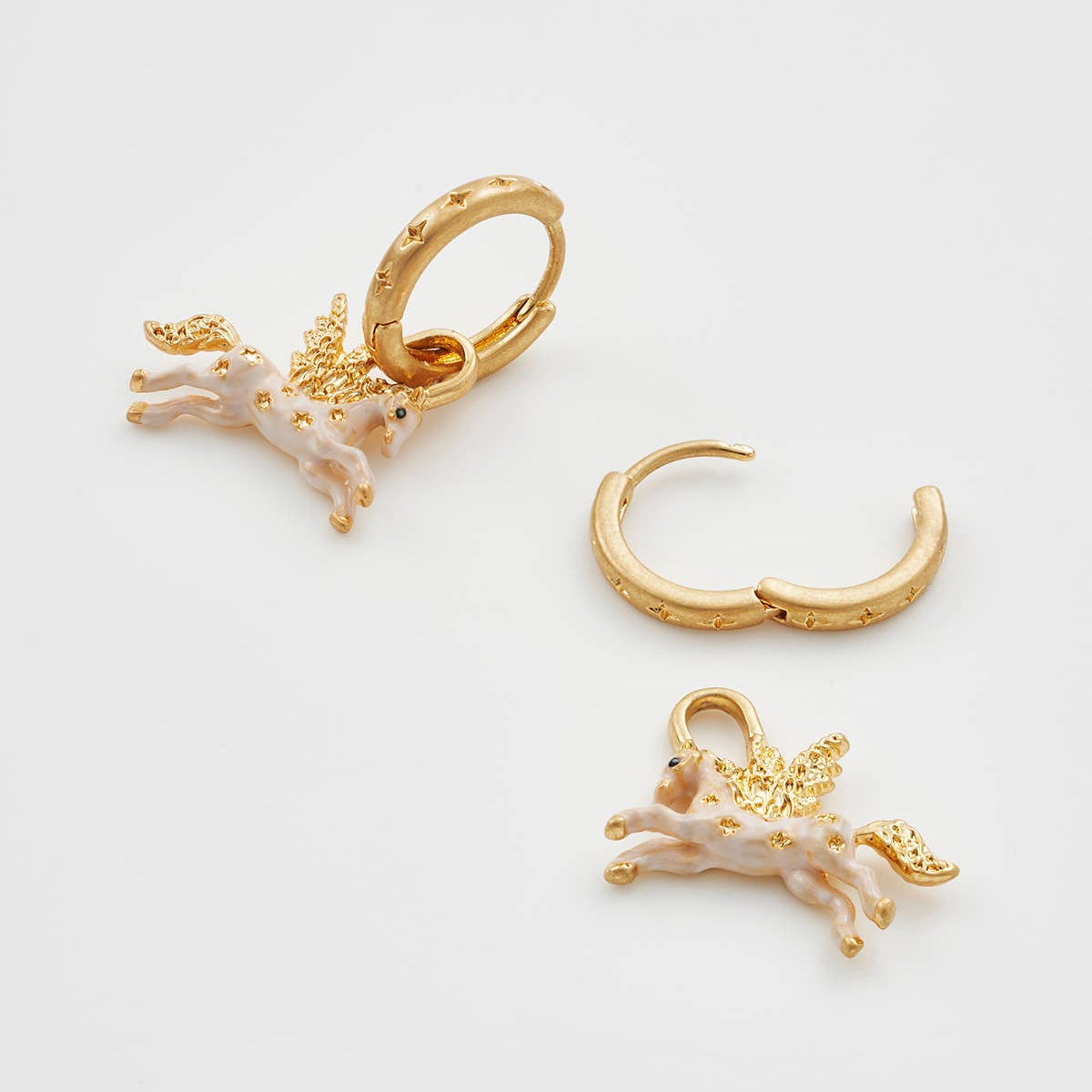 A set of gold-toned huggie hoop earrings and matching drop earrings, each featuring a small winged unicorn charm in pale pink enamel with gold detailing, displayed on a clean white background.