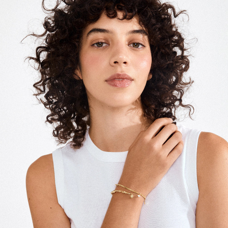 Woman with curly hair wearing a white sleeveless top and gold jewelry on a plain background