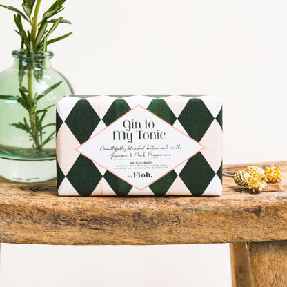 A rectangular bar of soap wrapped in a dark green and white argyle patterned paper sleeve with a white diamond label reading 'Gin to My Tonic' and 'by Floh.' It sits on a rustic wooden surface next to a glass vase with green stems and small dried flowers.