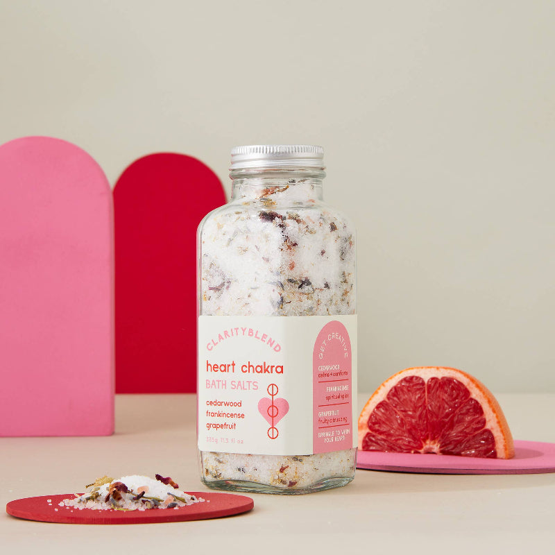 A glass bottle of Charity Blend Heart Chakra Bath Salts, filled with white and pink salts, is displayed on a beige surface with a pink and red background. A slice of grapefruit and a small dish of the bath salts are styled as props.