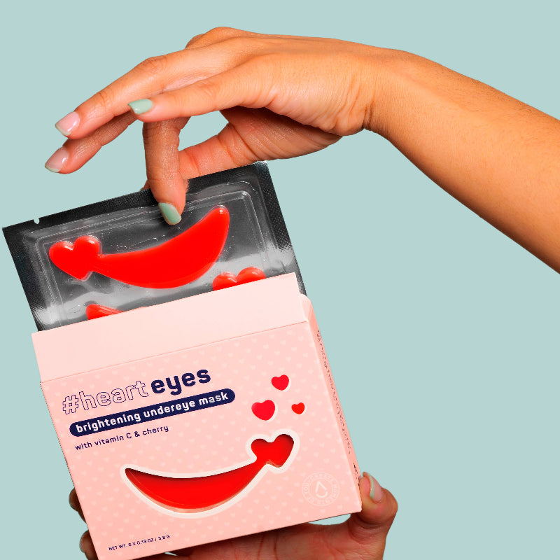 Hand holding a 'heart eyes' brightening under-eye mask against a light blue background