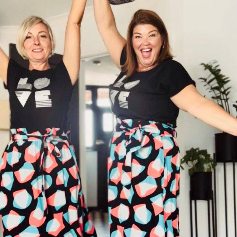 Two women wearing matching black t-shirts with white designs and colorful skirts, posing joyfully.