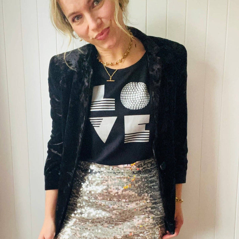 Woman wearing a black blazer, black 'LOVE' graphic shirt, and silver sequin skirt against a white wall.