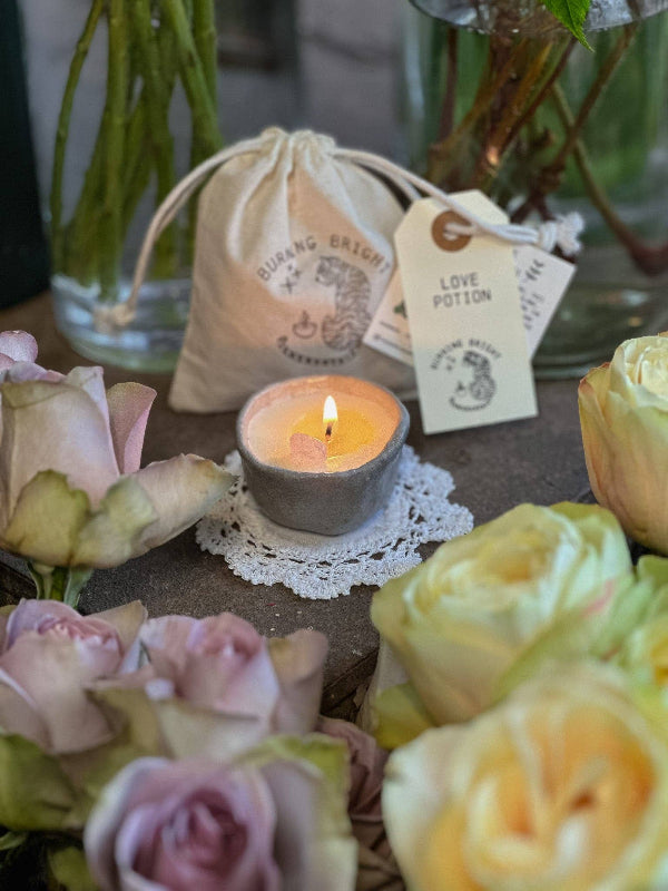 Candle with a decorative bag and tags surrounded by flowers on a table.
