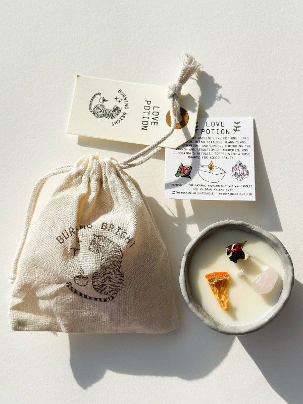 Candle with decorative stones in a small dish, surrounded by branded bags and cards on a light background