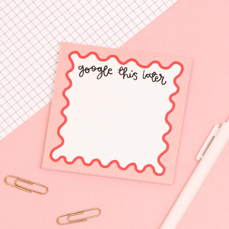 Notepad with 'Google this later' text on a pink surface with a pen and paper clips.