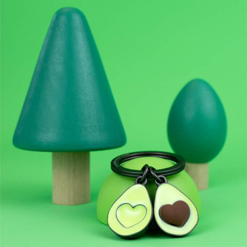 Two green avocado-halves on a keychain with heart designs on a green background with two small green trees.