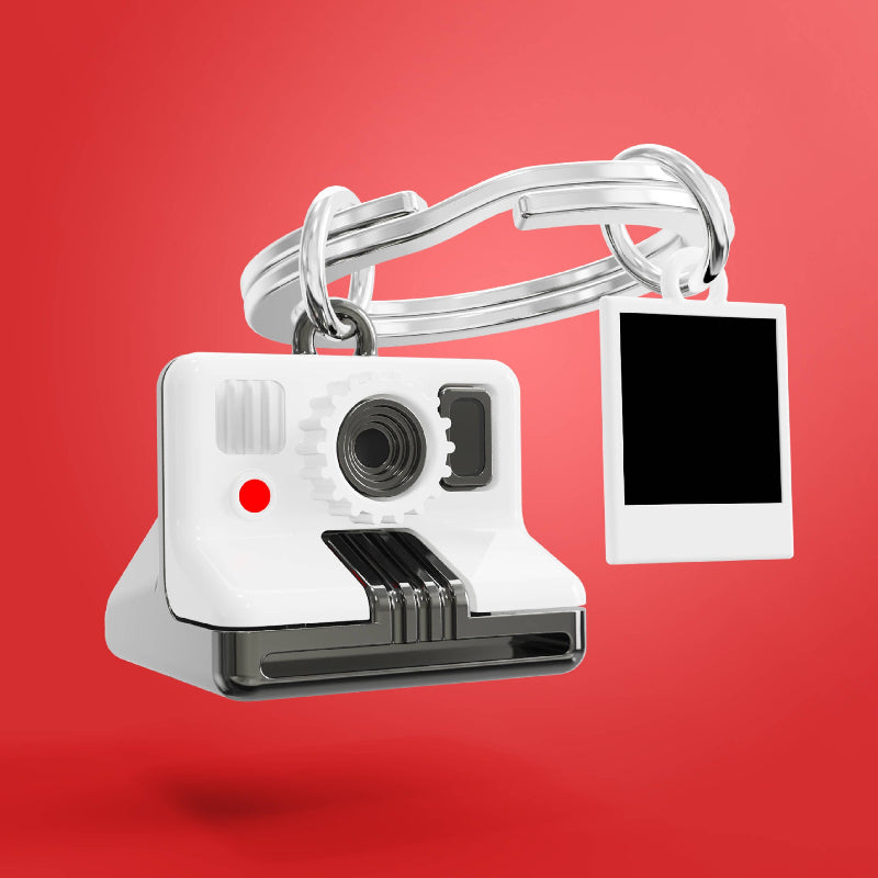 A novelty keychain featuring a white and silver instant camera design with a small red light, attached to a split ring and a white square frame that resembles a Polaroid photo holder, shown against a solid red background.