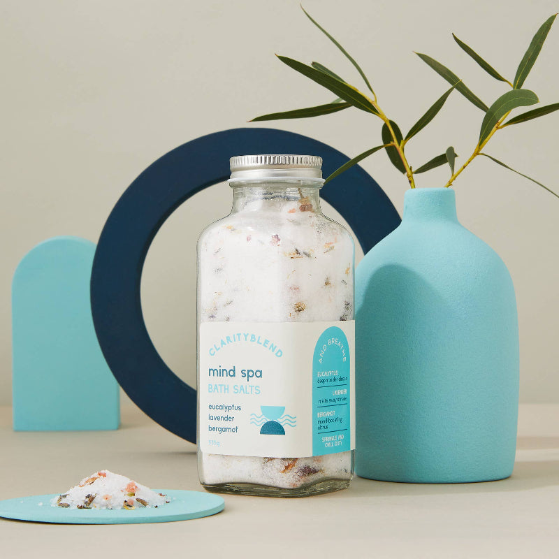 A glass jar of Charity Blend 'mind spa' bath salts sits on a light beige surface, styled with a light blue ceramic vase holding green leaves, a small dish with salt, and abstract blue geometric shapes against a neutral background.