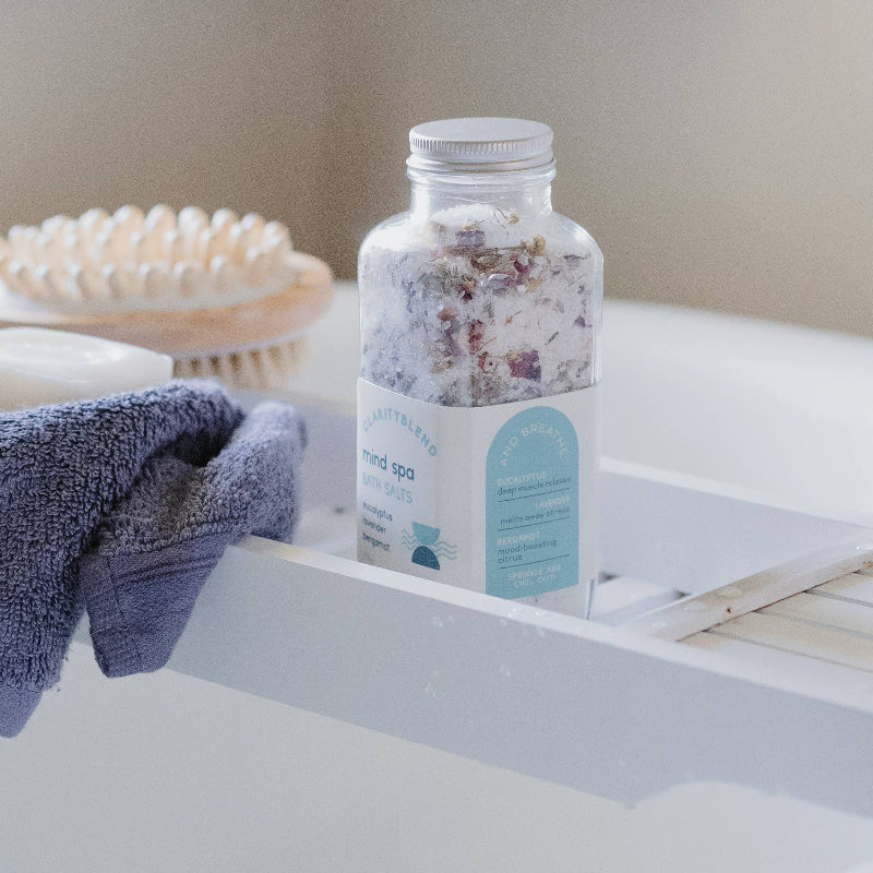 A clear bottle of Mind Spa mineral bath salts sits on a white bathtub ledge, with a folded navy towel and a wooden scalp massager in the background.