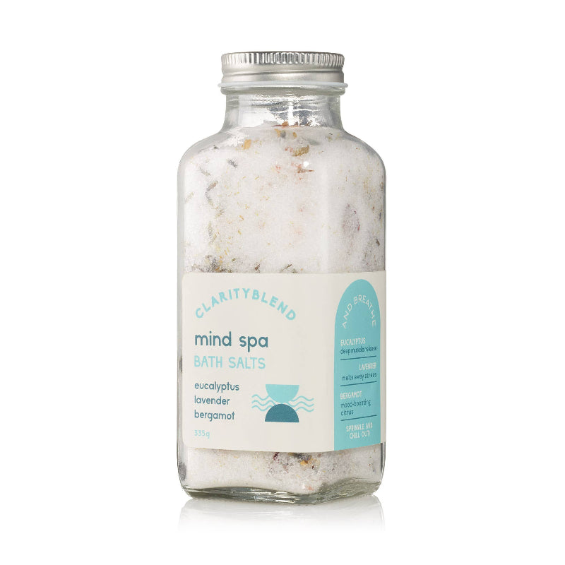 A glass jar of Clarity Blend Mind Spa Bath Salts, featuring white salt crystals with visible botanical pieces inside, a silver metal cap, and a white label with light blue text and graphics.