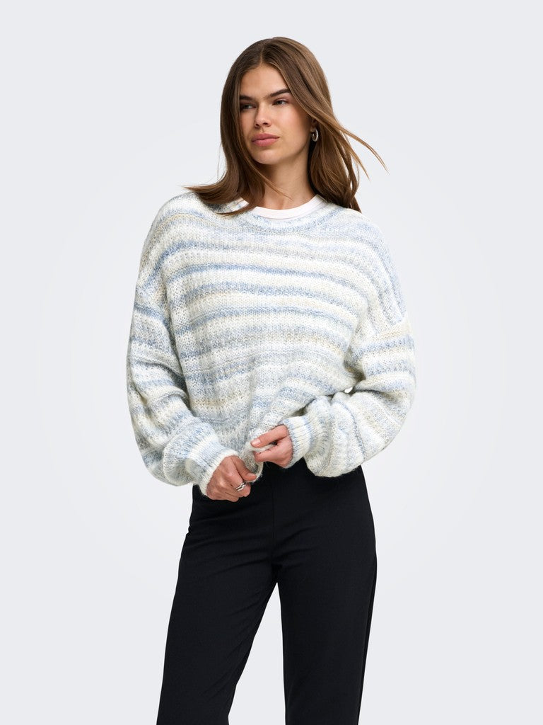A woman modeling a fuzzy, textured knit crewneck sweater with horizontal stripes in white, light blue, and beige, paired with black pants, against a solid light gray background.