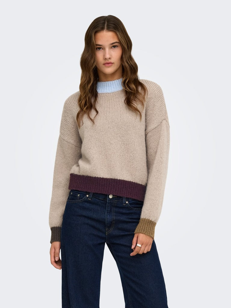A woman modeling a beige knit sweater with a contrasting dark purple ribbed hem and brown ribbed cuffs, paired with dark blue jeans, against a light gray studio background.