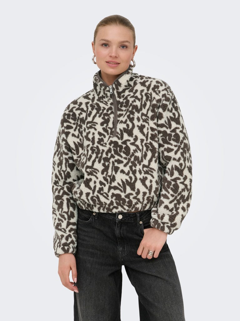 A woman modeling a beige and dark brown animal print quarter-zip fleece pullover, paired with dark gray jeans, against a light gray studio background.