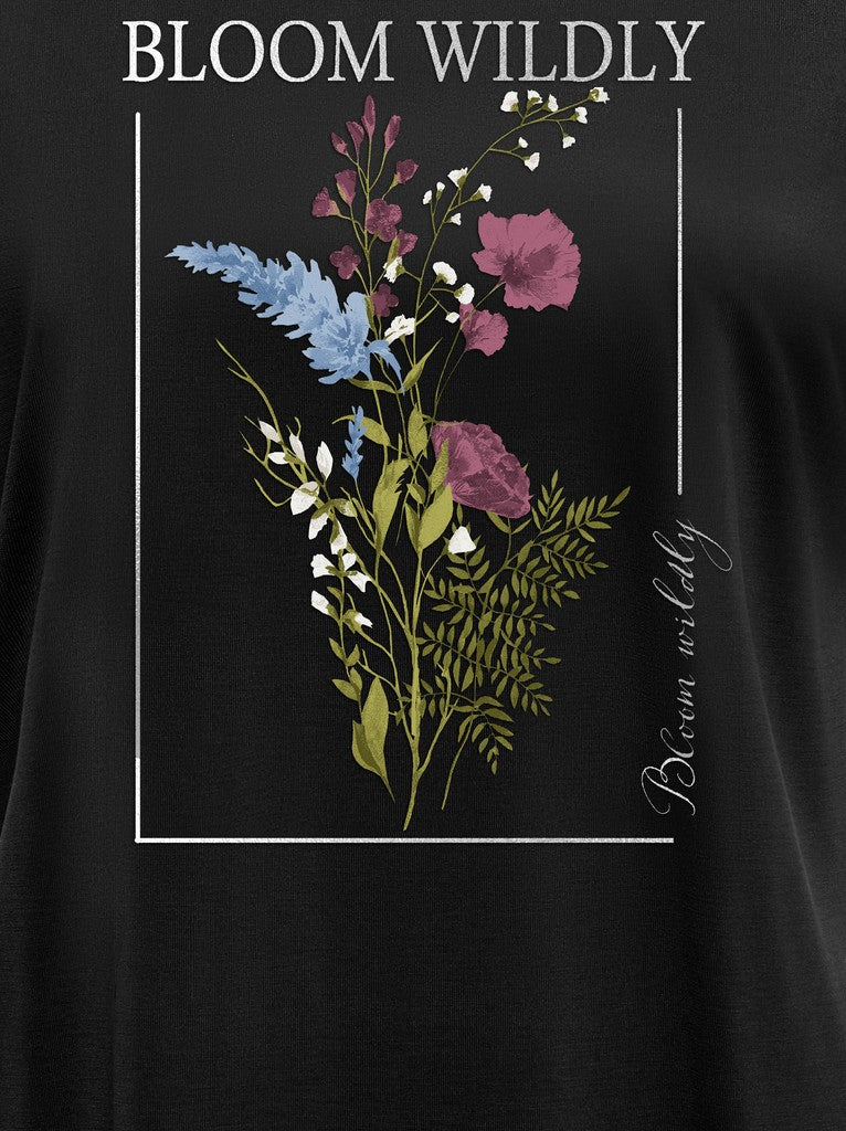A close-up of a black garment featuring a botanical graphic print with purple flowers, green leaves, and blue accents, framed by white text reading 'BLOOM WILDLY' and 'Bloom wildly' on a black background.