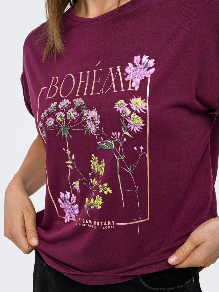 A woman wearing a burgundy short-sleeve graphic T-shirt with 'BOHÉM' in gold lettering and botanical illustrations in purple, yellow, and green, paired with black pants against a light gray background.