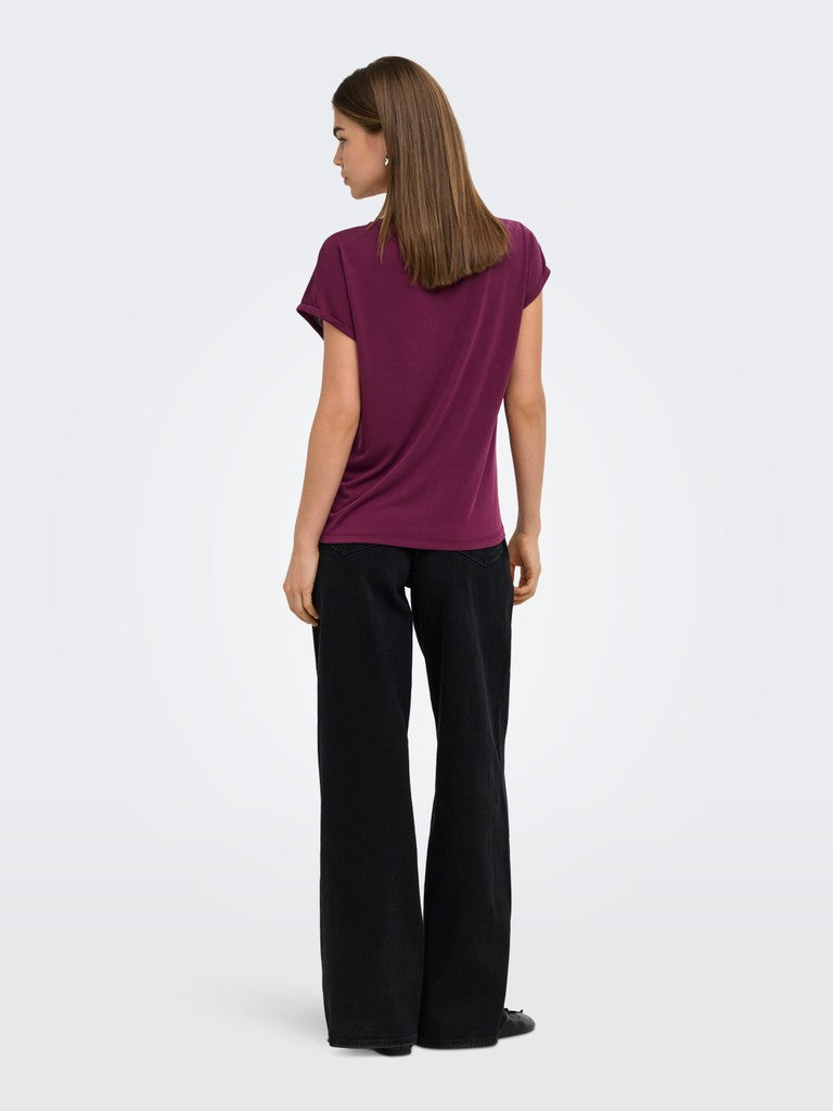 A woman viewed from the back wearing a short-sleeve deep plum-purple t-shirt and black wide-leg trousers, standing against a light gray studio background.