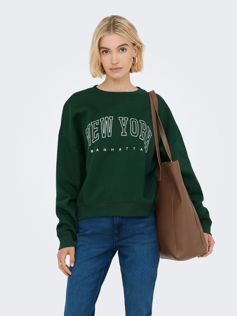 A woman modeling a dark green crewneck sweatshirt with 'NEW YORK MANHATTAN' printed in white across the chest, paired with blue jeans and a brown tote bag, against a light gray studio background.