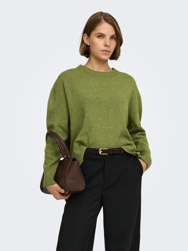 A woman modeling an olive green knit sweater with black trousers and a brown handbag, photographed against a light gray studio background.