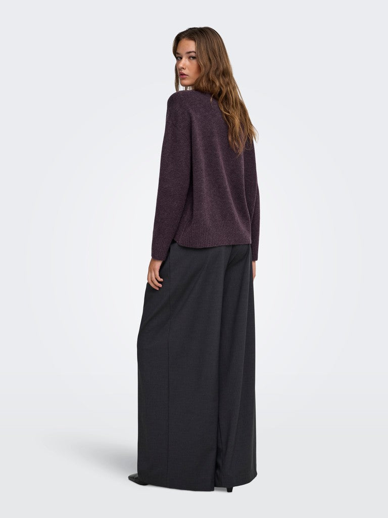 A woman is shown from the back wearing a dark plum-purple knit cardigan and wide-leg black trousers, standing against a light gray studio background.
