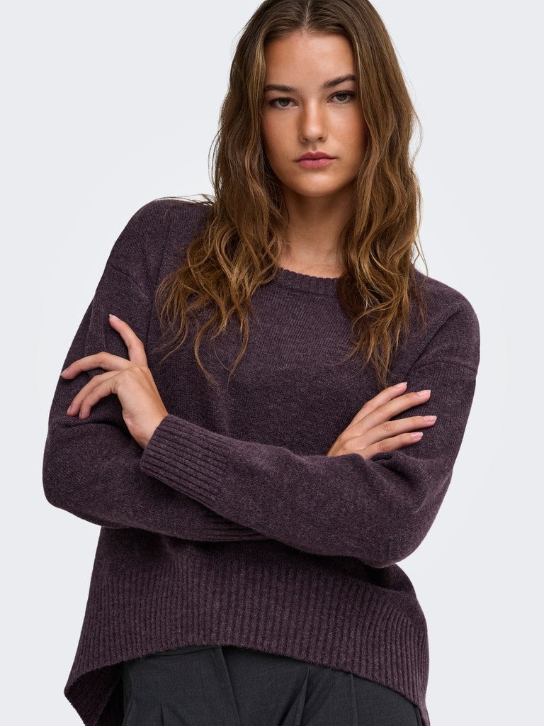 A woman with long wavy brown hair wearing a dark plum purple knit sweater with crossed arms, photographed against a light gray studio background.