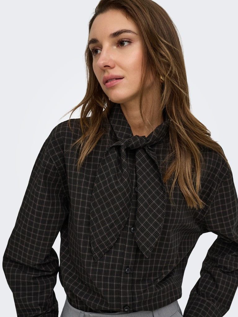 A woman wearing a black long-sleeve button-up shirt with a subtle windowpane plaid pattern and a large bow tie collar, photographed against a clean light gray studio background.