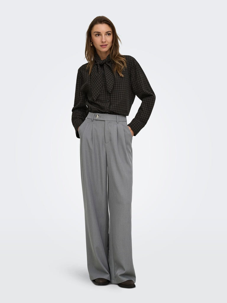 A woman modeling a black patterned long-sleeve blouse with a bow tie collar and high-waisted, wide-leg gray trousers against a light gray studio background.