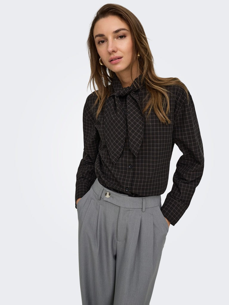 A woman modeling a black long-sleeve button-up shirt with a subtle windowpane plaid pattern and a ruffled tie-neck collar, paired with gray pleated trousers, against a light gray studio background.