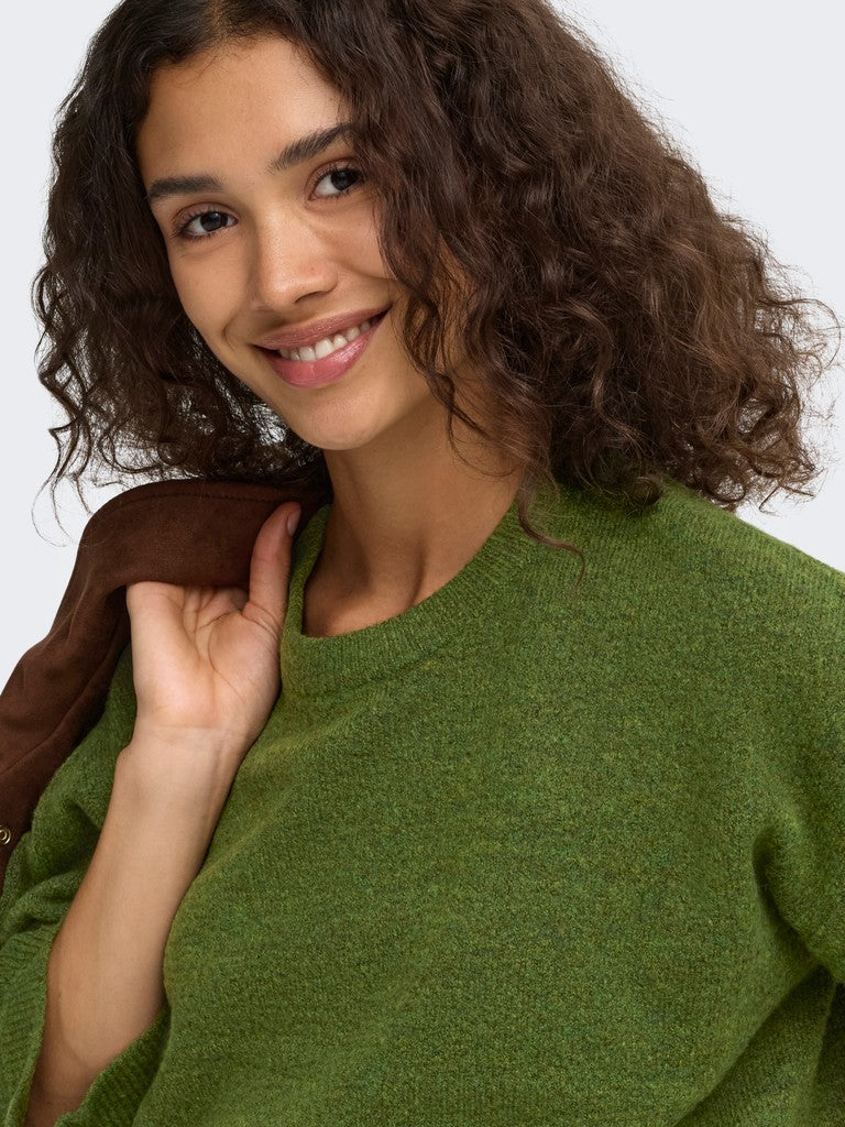A close-up of a smiling woman with curly brown hair wearing a textured olive green knit sweater and holding a brown jacket over her shoulder, set against a plain light gray background.