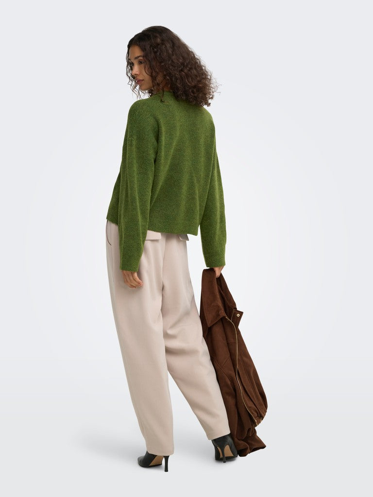 A woman is shown from the back wearing a fuzzy olive green cropped sweater, cream wide-leg trousers, and black heels, while holding a brown coat against a plain light gray studio background.