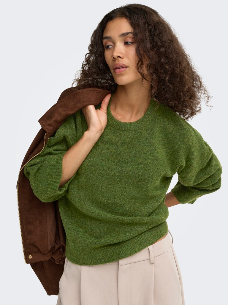 A woman with curly brown hair wearing a textured olive green crewneck sweater, a brown jacket draped over her shoulder, and light beige trousers, photographed against a light gray studio background.