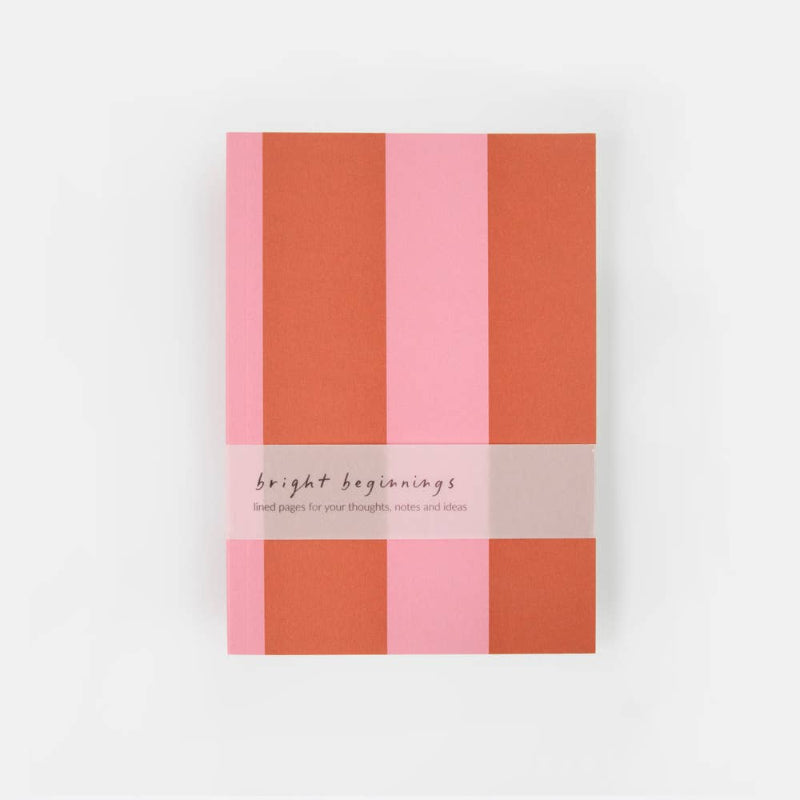 A notebook with a striped cover in shades of pink and terracotta orange, wrapped with a white paper band that reads 'bright beginnings'.