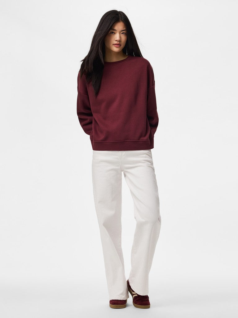 A woman modeling a dark burgundy crewneck sweatshirt and off-white straight-leg pants against a plain white studio background.