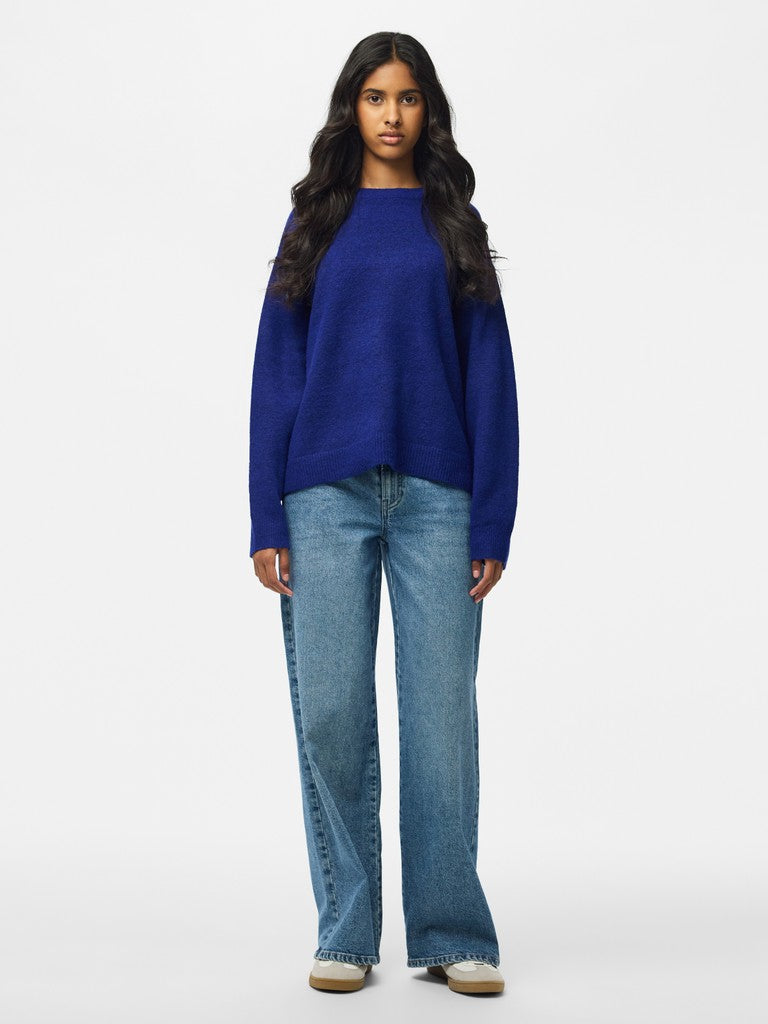 A woman modeling a vibrant royal blue knit sweater with wide legs, paired with light wash blue jeans and white sneakers, set against a plain white studio background.