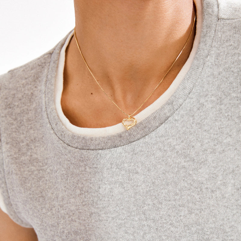 Close up of someone wearing a gold heart necklace on a grey shirt on a white background