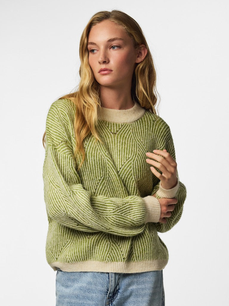 A woman wearing a light olive green cable-knit sweater with beige ribbed trim, paired with blue jeans, against a plain light gray background.