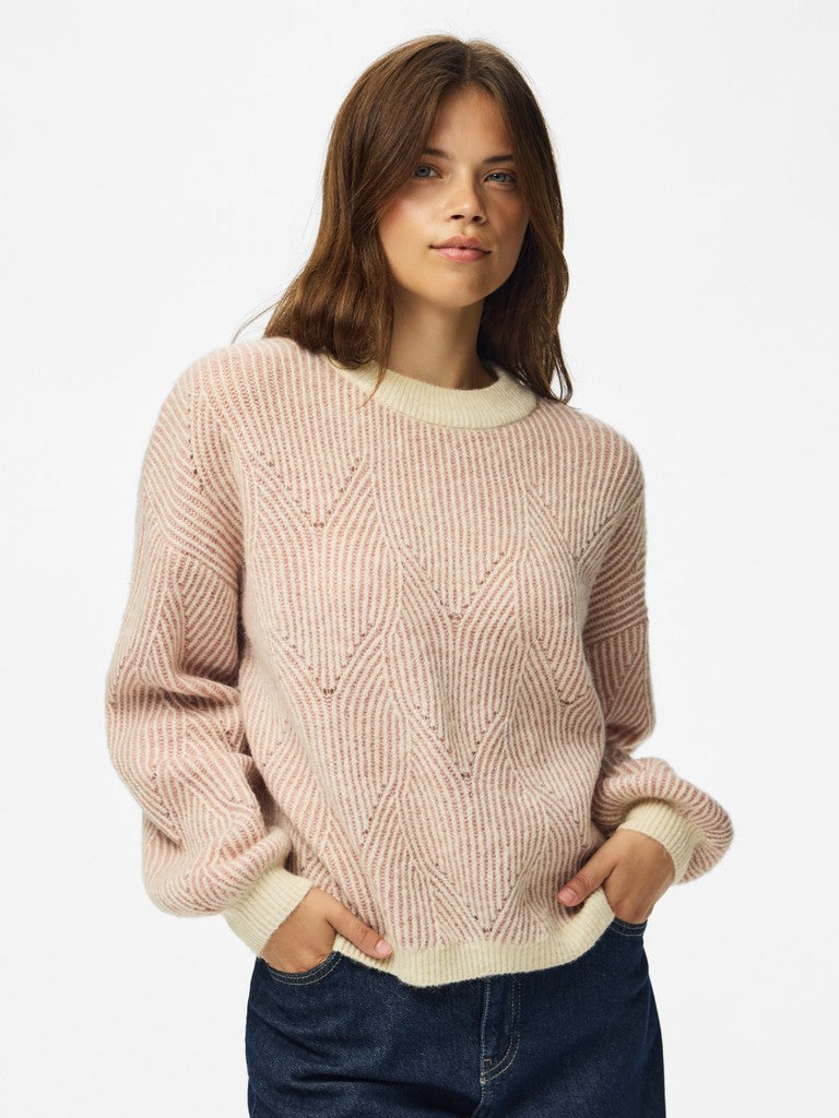 A woman wearing a light pink cable-knit crewneck sweater with cream ribbed trim, paired with dark blue jeans, against a plain white studio background.