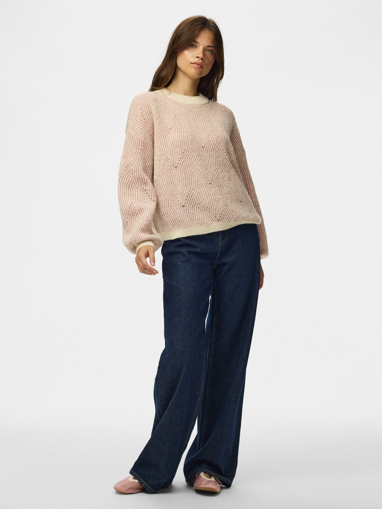 A woman modeling a light pink knit crewneck sweater with long sleeves and dark blue wide-leg jeans, standing against a plain light gray studio background.