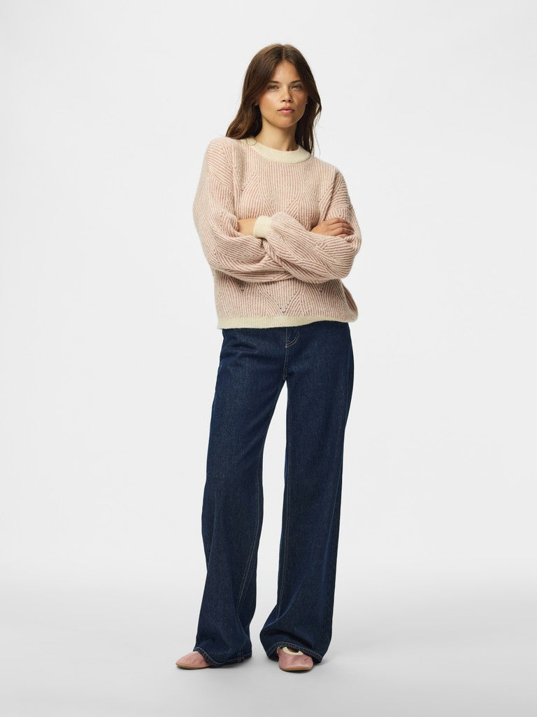A woman stands against a light gray studio background wearing a loose, oversized knit sweater in a pale blush pink and dark blue wide-leg jeans.